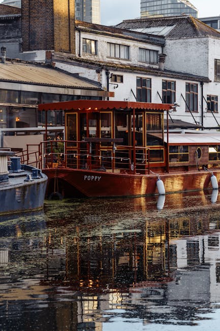 A waterfront scene in Little Venice featuring two narrowboats docked along the canal, with a brick apartment building in the background. The canal water reflects the boats and greenery, including a ta