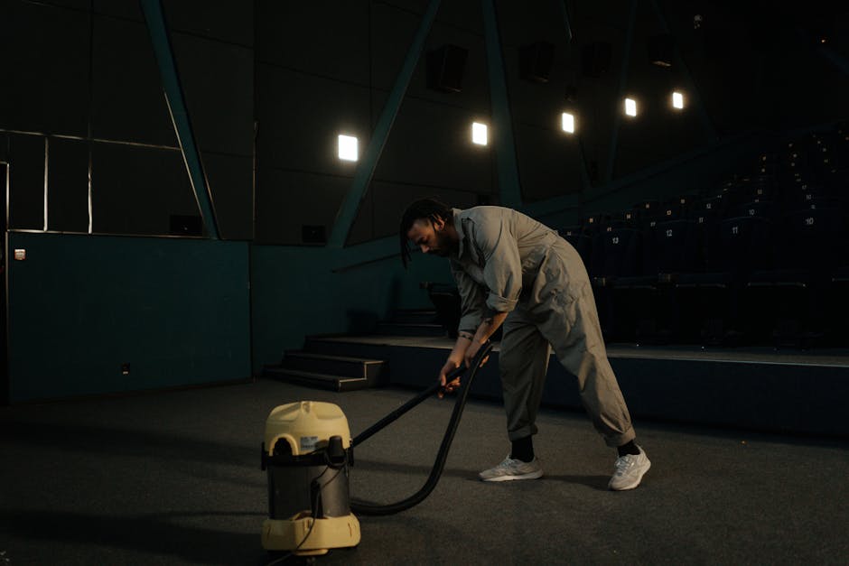A person is vacuuming a traditional patterned area rug with a handheld vacuum cleaner in a residential setting. The rug has floral motifs in beige, green, and orange shades, covering a wooden floor. T