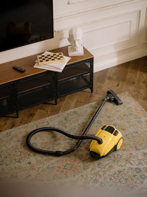 A yellow canister vacuum cleaner with a black flexible hose and a floor attachment is positioned on a patterned area rug in a residential living room. In the background, there is a black metal media console with a wooden top, holding a remote control, a stack of books, and a decorative white bust sculpture. The room features light-colored wooden flooring, white paneled walls, and a large flat-screen TV mounted on the wall above the console. Natural light illuminates the space, highlighting the clean and well-maintained surfaces, suitable for surface cleaning and deep cleaning assessments in a domestic setting as part of routine hygiene and maintenance by Paddington Carpet Cleaners.